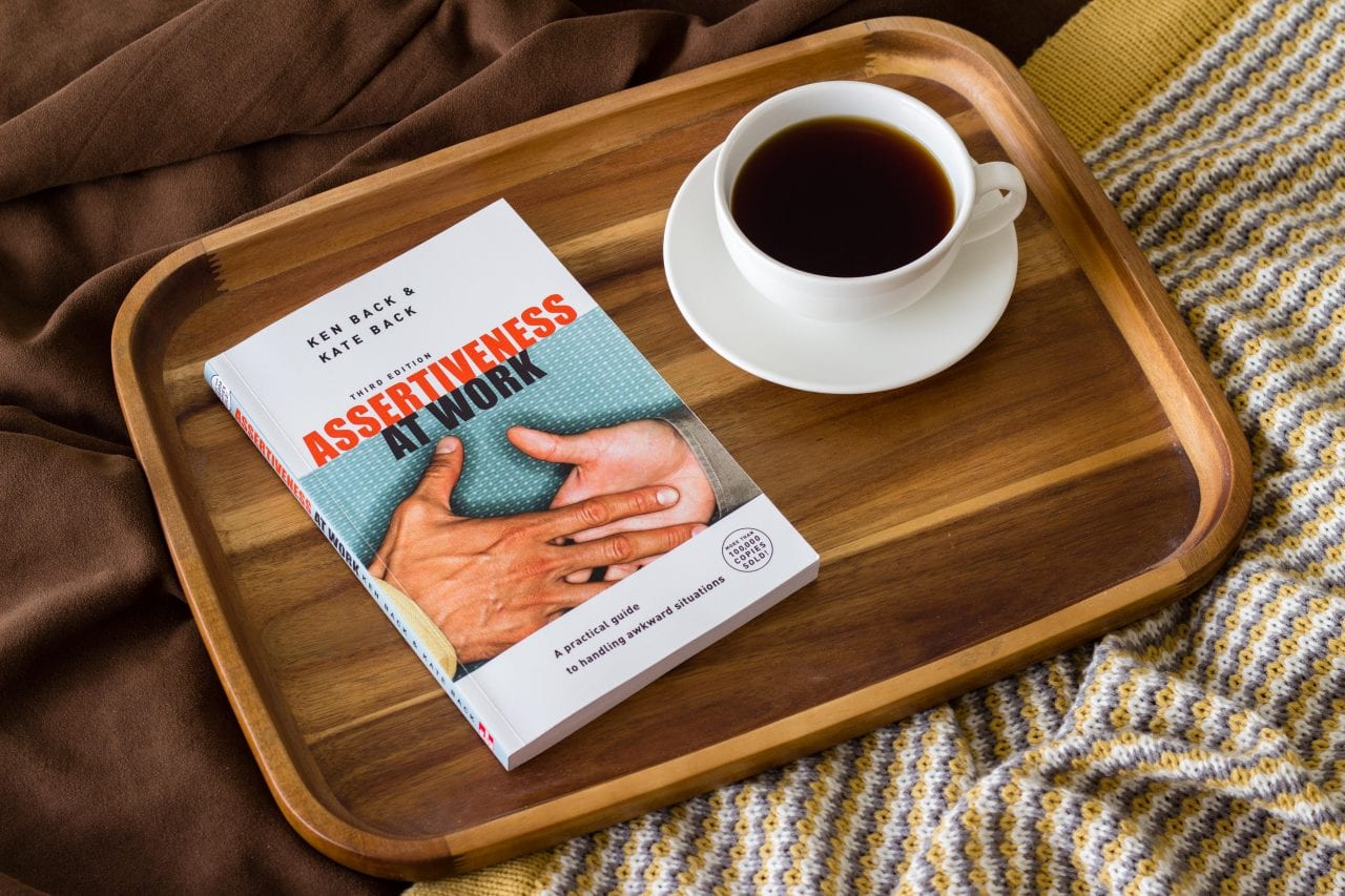 The book "Assertiveness at Work" and a cup of coffee on a wooden tray