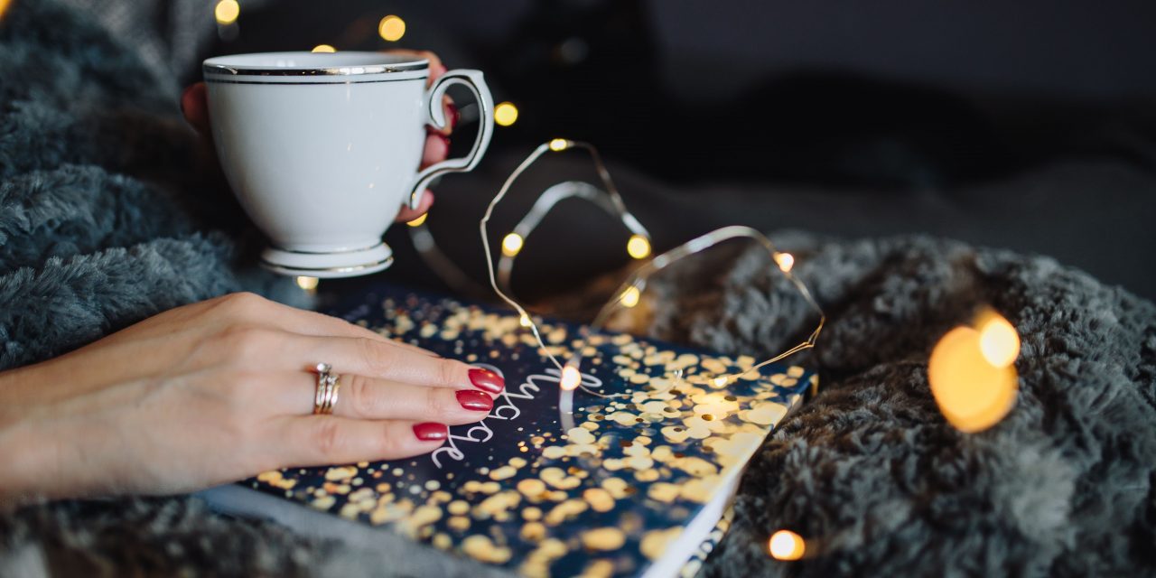 A fair-skinned woman holding a cup of tea and reading a book entitled "Hygge"