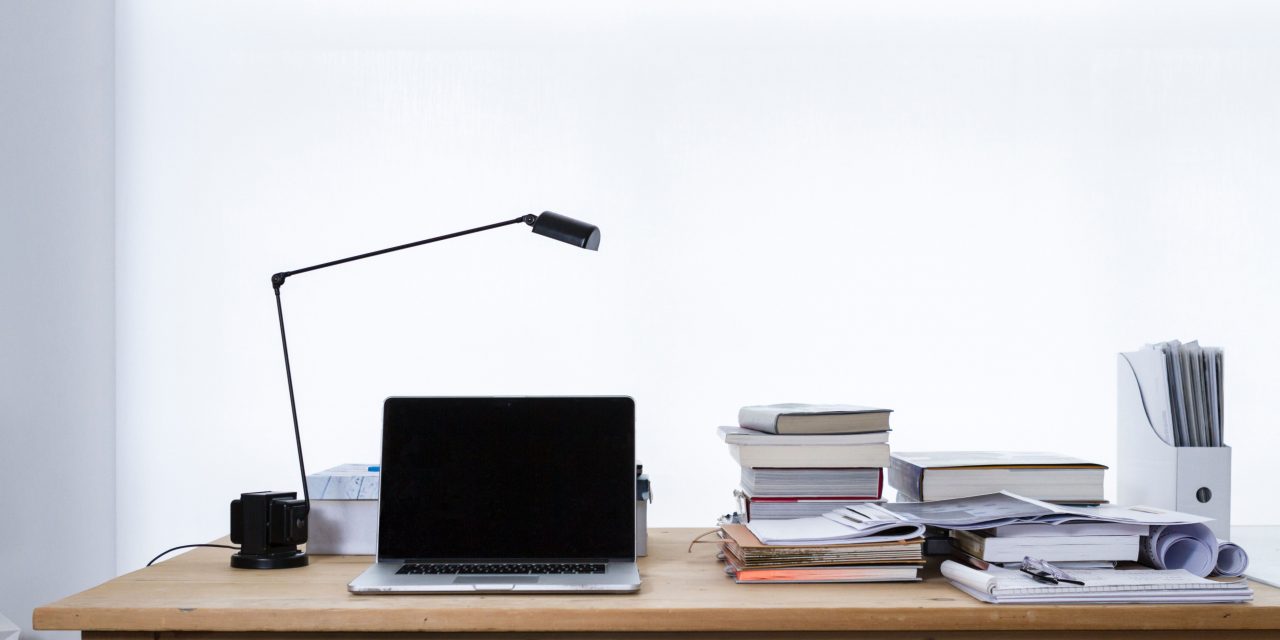 A laptop and several piles of paper and books on a wooden desk