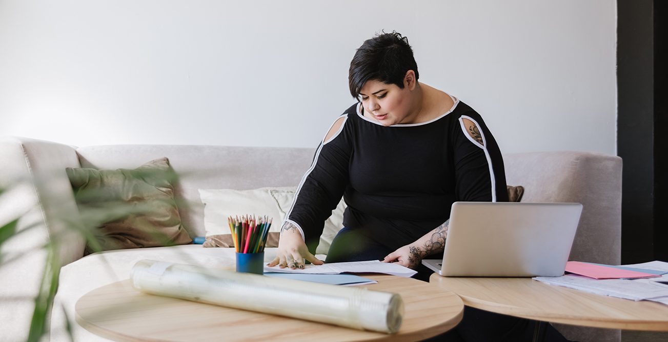 A woman sitting on a couch, with her laptop on the coffee table, looking at some papers