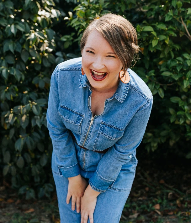 Christina is wearing a denim boiler suit and is doubled over in delighted laughter.