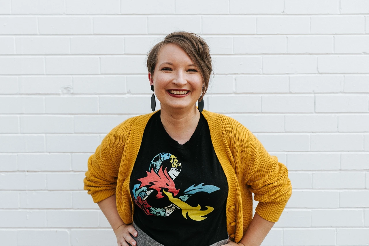 Christina is standing in front of a white brick wall, wearing a bright yellow cardi and a wide, eye-crinkling smile. Her hands are on her hips.
