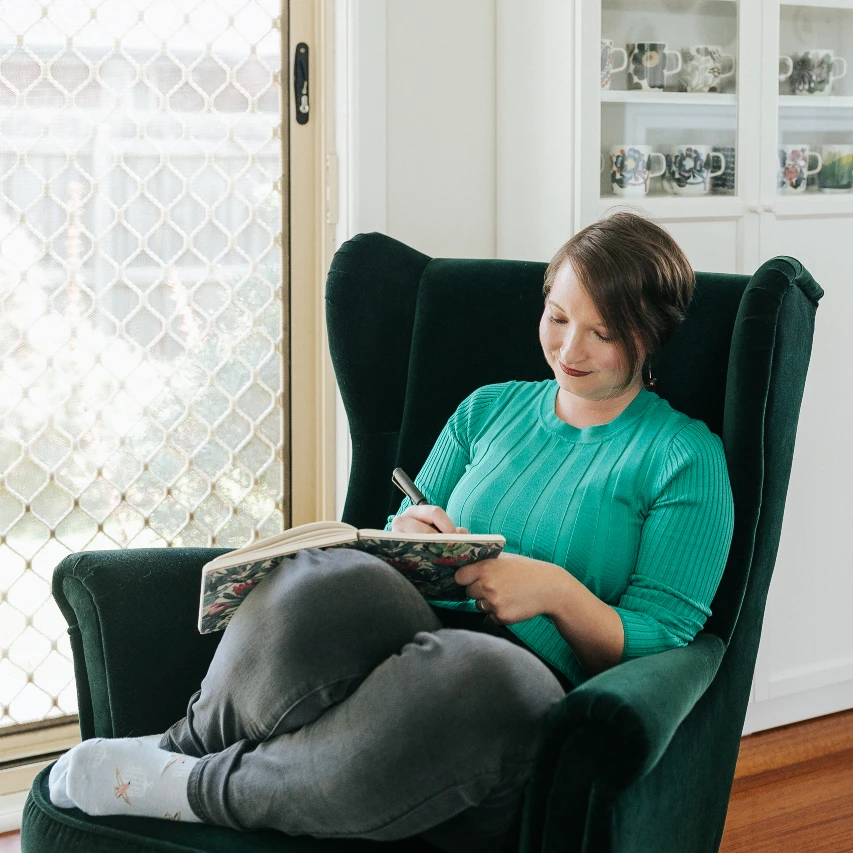 Christina is sitting in a green velvet wingback chair, with her legs tucked up, writing in a notebook