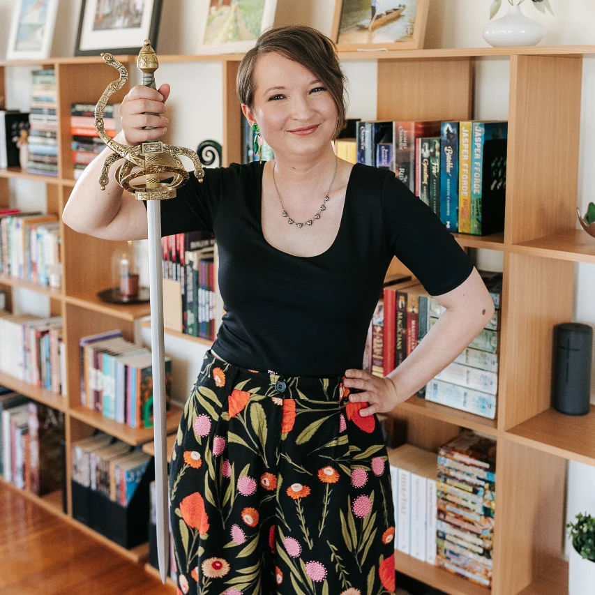 Christina is standing in front of a bookshelf, holding a plastic prop sword and smiling