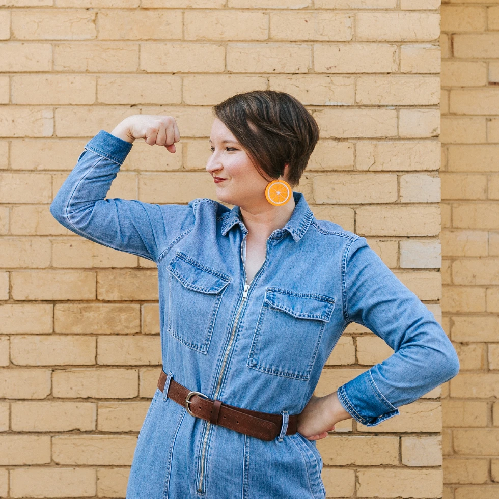 Christina is wearing a denim boiler suit and striking a muscle-flexing pose, despite having no muscles to speak of.
