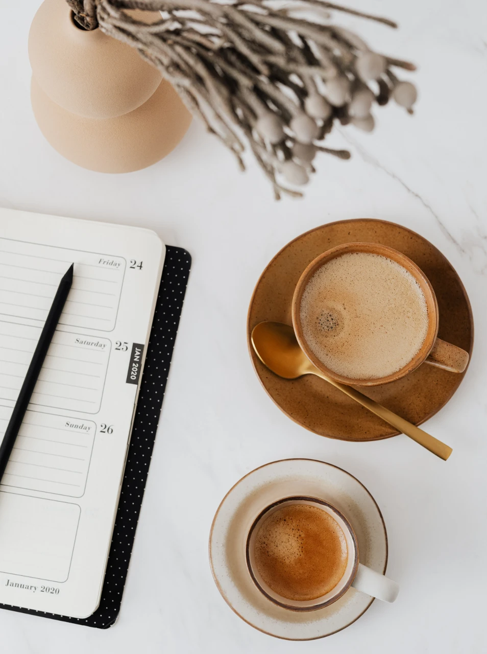 Two cups of coffee sit on a marble table, beside an open planner