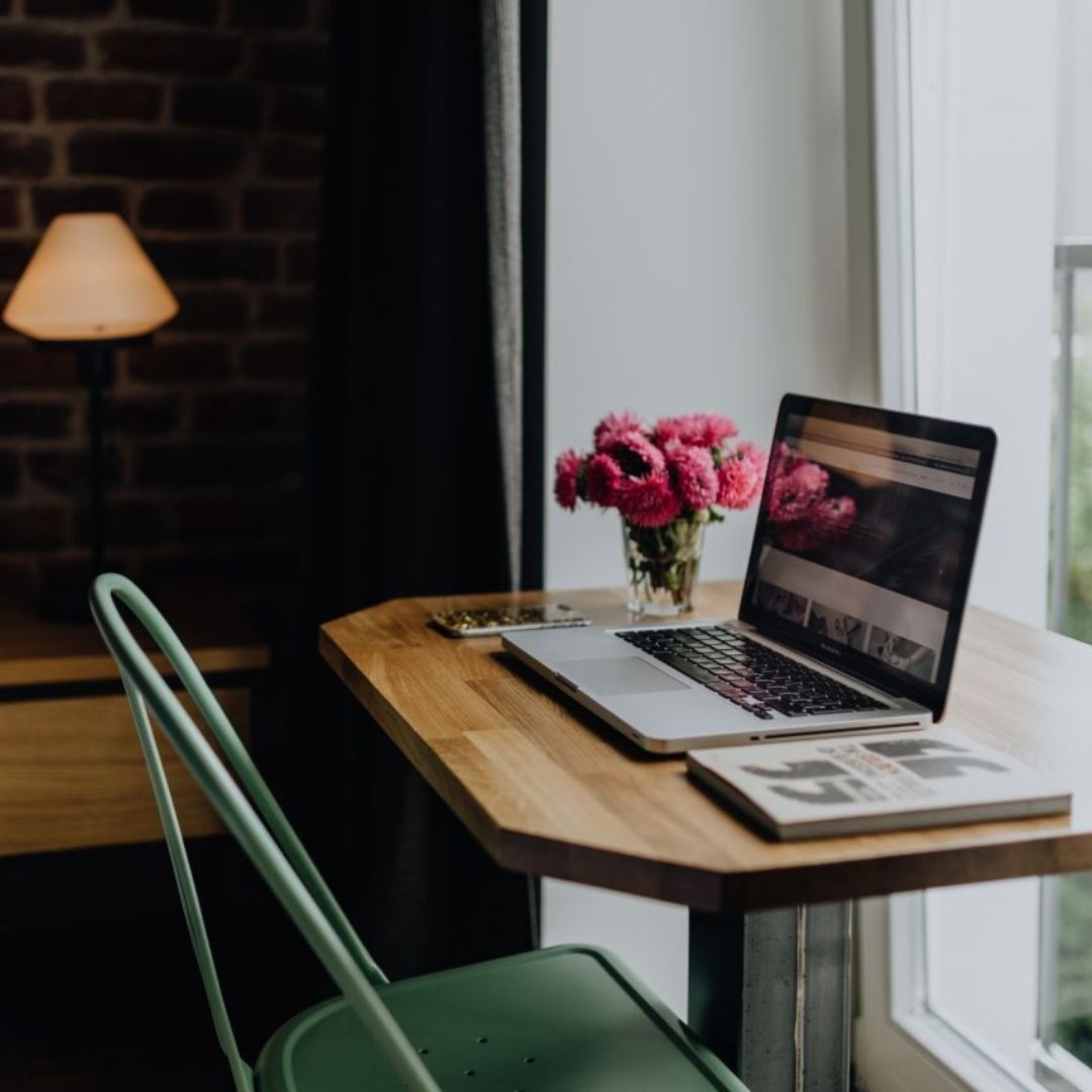 A laptop sits on a small desk by a window, in a bedroom