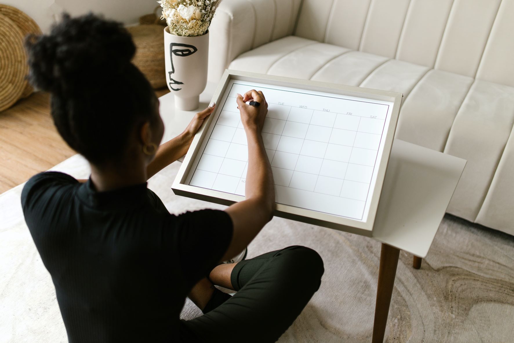 A brown-skinned woman is sitting on the floor, writing on a whiteboard calendar. Her hair is up in a curly bun.