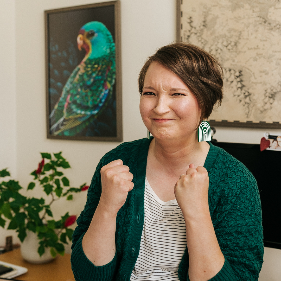 Christina is wearing a very frustrated expression and is clenching her fists. She's wearing a cozy forest green cardigan and big green rainbow earrings.