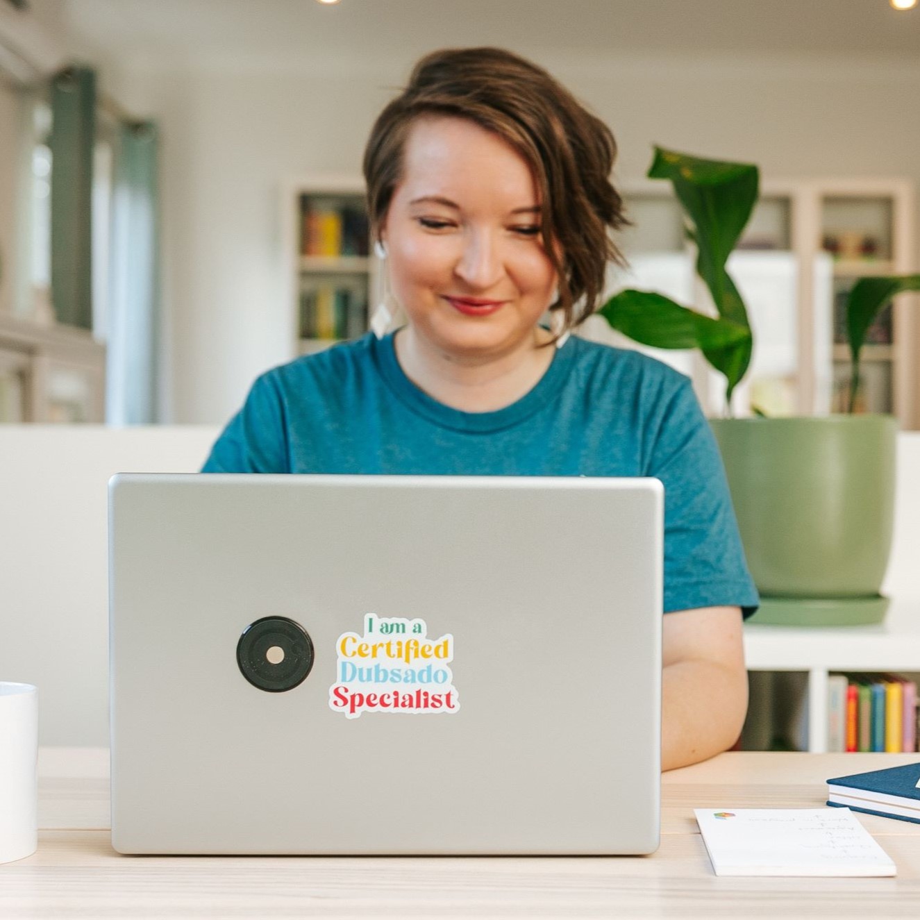 Christina is wearing a simple blue t-shirt and smiling while working on her laptop. The laptop has a sticker on it thaty says "I am a Certified Dubsado Specialist."