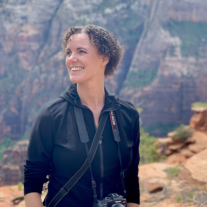 Rachel is standing before a backdrop of mountains, camera slung over her shoulder, smiling widely while she scopes out her hiking trail.