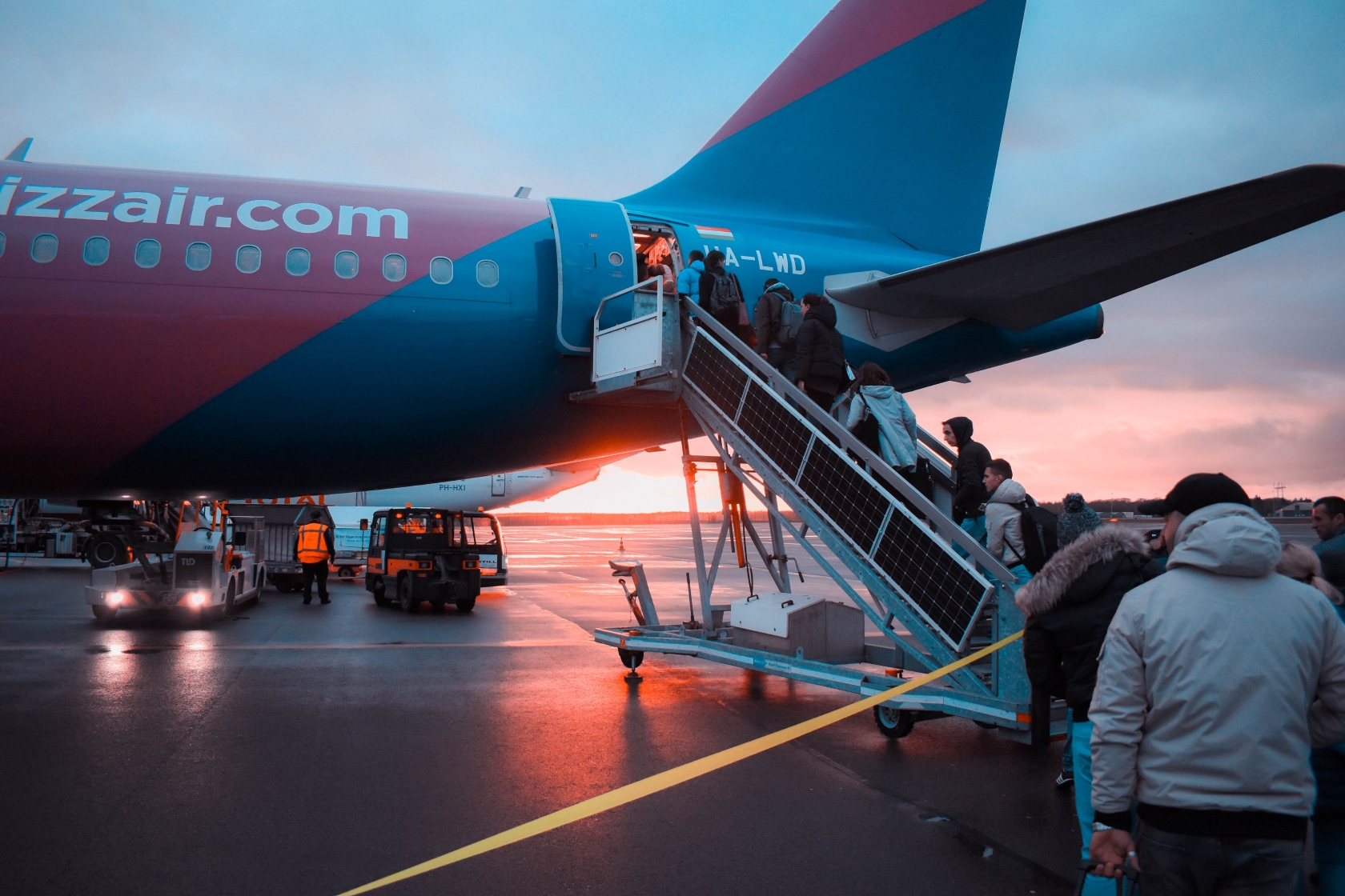 The back end of an aeroplane, on the tarmac at dusk. Portable stairs lead up to the plane's rear door and a group of people are queued up to get on board.