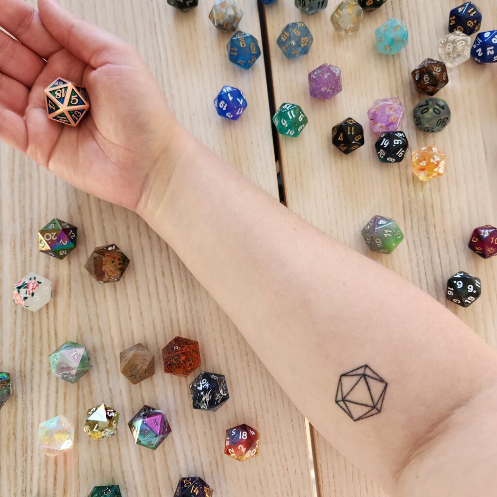 Christina's forearm, featuring a tattoo which shows the outline of a 20-sided polyhedral shape. The formarm is resting on a table, surrounded by 20-sided dice.